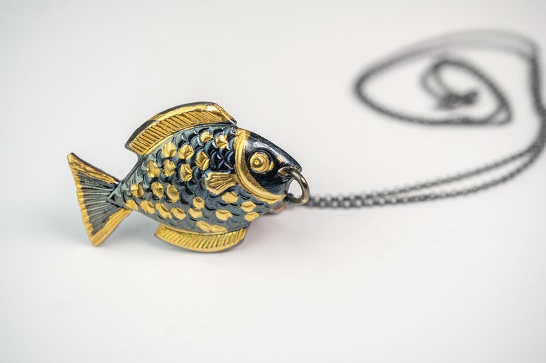 K&ouml;nnte beinhalten: Eine Halskette mit einem goldenen und schwarzen Fisch-Anh&auml;nger an einer schwarzen Kette. Der Fisch ist detailliert mit Schuppen und einem kleinen Auge.