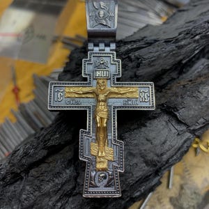 Könnte beinhalten: Ein orthodoxer Kreuzanhänger aus Silber und Gold mit filigranen Details. Das Kreuz zeigt auf der Vorderseite eine Darstellung von Jesus Christus und oben die Inschrift "IC XC NIKA".