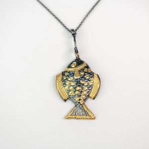 K&ouml;nnte beinhalten: Eine silberne Kettenkette mit einem goldenen und schwarzen Fisch-Anh&auml;nger. Der Fisch hat ein detailliertes Design mit Schuppen und Flossen.