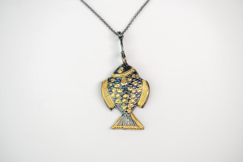 K&ouml;nnte beinhalten: Eine silberne Kettenkette mit einem goldenen und schwarzen Fisch-Anh&auml;nger. Der Fisch hat ein detailliertes Design mit Schuppen und Flossen.