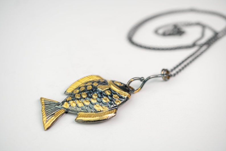 K&ouml;nnte beinhalten: Eine Halskette mit einem goldenen und schwarzen Fisch-Anh&auml;nger an einer zarten Kette. Der Fisch ist mit Schuppen verziert und hat einen einfachen Hakenverschluss.
