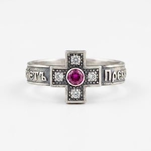 Peut inclure: Une bague en argent avec un motif central en forme de croix orné d'une pierre précieuse ronde rose foncé au centre, entourée de quatre petites pierres claires. L'anneau porte une inscription en cyrillique sur un fond sombre et oxydé.