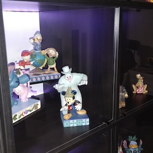 Op de afbeelding: Een zwarte glazen vitrinekast met planken vol met verschillende Disney-figuren. De figuren zijn gemaakt van beschilderd keramiek en bevatten personages zoals Mickey Mouse, Donald Duck, Goofy en Stitch. De kast is verlicht met paarse LED-verlichting.