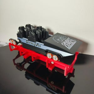May include: A miniature roller coaster car model featuring a black and light blue body with the text Worlds of Fun and Orient Express printed on the side and top. The model sits on a bright red track structure with orange-rimmed wheels.