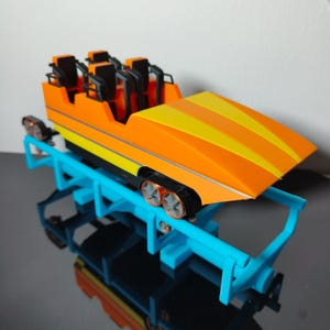 Puede incluir: Vagón de montaña rusa naranja y amarillo con asientos y ruedas negros. El vagón está en una vía azul.