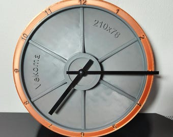 Puede incluir: Un reloj gris con un borde de color cobre y agujas negras. La esfera del reloj tiene seis radios y los números del 1 al 12 están impresos en el borde. El texto "210x76" y "Vekoma" están impresos en la esfera del reloj.