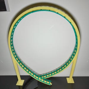 Pode incluir: Um loop de montanha-russa modelo amarelo e verde com uma pista que sobe e desce. O loop é suportado por dois postes amarelos.