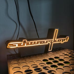 May include: A black and white illuminated sign with the word "schwarzkopf" in white letters on a black background. The sign is sitting on a black surface with a grid pattern.