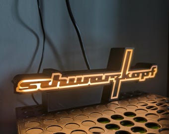 Puede incluir: Un letrero iluminado en blanco y negro con la palabra "schwarzkopf" en letras blancas sobre fondo negro. El letrero está colocado sobre una superficie negra con un patrón de cuadrícula.