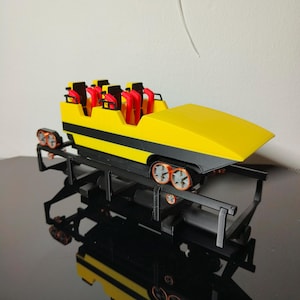 May include: A yellow and black roller coaster car with red seats. The car is on a black track and has orange wheels.