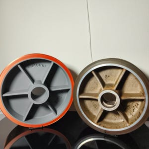 Op de afbeelding: Twee industriële wielen, een met een rode rubberen band en een grijs metalen hart, en de andere met een bruin metalen hart en een zilveren velg. Het wiel met de rode band heeft het woord "Tekoma" op het hart gedrukt.