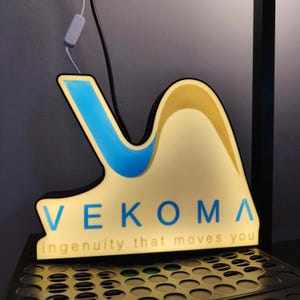 Può includere: Un logo 3D bianco e blu con il testo "VEKOMA ingenuity that moves you". Il logo ha la forma di un'onda ed è illuminato da dietro.