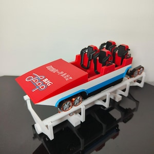 May include: A miniature roller coaster model with a red, white, and blue color scheme. The model features a red car with black seats, a white track, and the words "BIG LOOP" and "HEIDE-PARK" printed on the front.