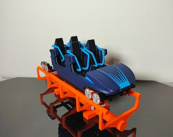 May include: A miniature roller coaster car model featuring a dark blue body with bright blue accents, black seats, and light blue safety restraints. The car is mounted on a vibrant orange track structure with visible silver wheels.