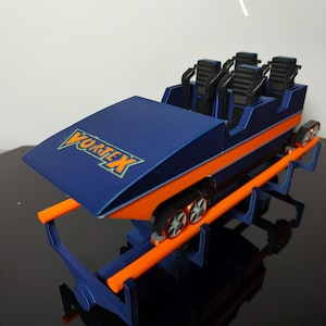 Op de afbeelding: Een blauwe en oranje model achtbaan trein met het woord "Vortex" erop gedrukt. De trein heeft zes zitplaatsen en staat op een baan.