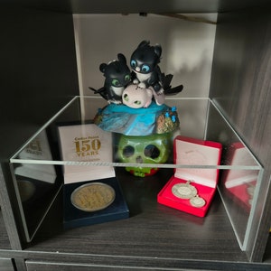 May include: A clear acrylic display case containing a black and green skull, a small pink figurine, and two black figurines. The case also features a gold coin with the text "150 Years" and a red box with two silver medals.