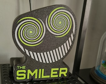 May include: A black, textured, shaped sign featuring two lime green and white spiral eyes and a wide, white-striped grin. The text THE SMILER is printed in lime green at the bottom of the sign, which is constructed from a dark, granular material.