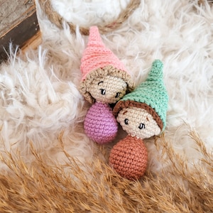 Puede incluir: Tres gnomos de crochet con sombreros y cuerpos de diferentes colores. Los gnomos están sentados sobre una superficie blanca y esponjosa con una planta de hierba seca marrón en primer plano.