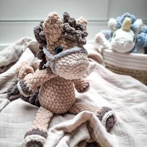 Puede incluir: Un caballo de ganchillo hecho a mano en tonos beige y marrón, con un ojo azul y una brida gris. El peluche tiene una melena y cola esponjosas, y está sentado sobre una manta suave de color neutro. Un unicornio de ganchillo está en el fondo.