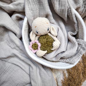 May include: A crocheted turtle toy with a pink flower, sitting in a white basket on a grey fabric background.