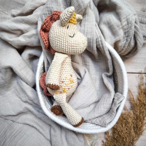 Può includere: Un giocattolo di unicorno all'uncinetto con un corpo marrone chiaro, una criniera rosa e un corno giallo. Il giocattolo è sdraiato in un cesto intrecciato bianco su uno sfondo di tessuto grigio.