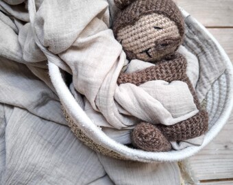May include: A brown crocheted sloth toy is nestled in a white and beige woven basket lined with a light beige muslin fabric.