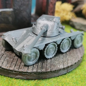 以下が含まれることがあります： グレーのミニチュア戦車のモデル。砲塔が持ち上げられ、4つの車輪が付いています。戦車は木目調の円形の台座の上に置かれています。