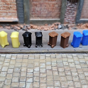 Könnte beinhalten: Eine Sammlung von Miniatur-Mülltonnen in verschiedenen Farben, darunter Gelb, Schwarz, Braun und Blau. Die Tonnen sind auf einer Pflasteroberfläche angeordnet, was eine Straßenszene suggeriert. Die Tonnen haben Deckel und Räder.