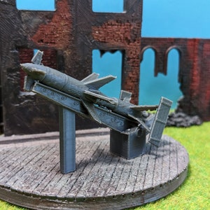 Op de afbeelding: Een grijs, 3D-geprint model van een raket op een standaard. De raket is beschadigd en er missen een paar onderdelen. De standaard is gemaakt van grijs, getextureerd materiaal. Het model is geplaatst tegen een achtergrond van een bakstenen gebouw.