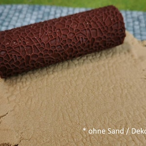Könnte beinhalten: Eine braune, strukturierte Rolle wird verwendet, um ein Muster in einem hellbraunen sandartigen Material zu erstellen. Der Text *ohne Sand / Deko* ist in der unteren rechten Ecke des Bildes sichtbar.
