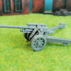 Könnte beinhalten: Ein graues Modell einer Artilleriekanone mit langem Lauf, Rädern und detailliertem Wagen. Die Miniaturkanone steht auf einer grünen Oberfläche, mit einem Hintergrund aus einem Backsteingebäude und Felsen.