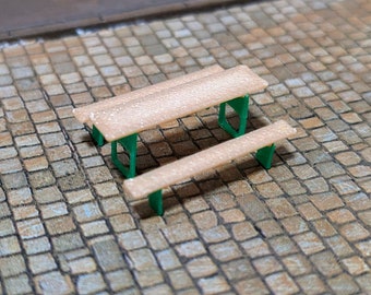 May include: A miniature wooden picnic table and bench set with green metal legs. The table and bench are both light brown and have a rough texture.