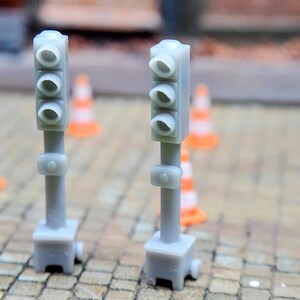 Pode incluir: Dois semáforos cinzentos em miniatura com três aberturas circulares cada. Vários pequenos cones de trânsito laranja e branco estão espalhados no fundo, sugerindo uma cena de construção ou modelismo.