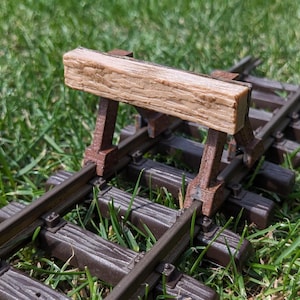 May include: A model railway track section with a wooden buffer stop. The track is brown and set on green grass. The buffer is made of light brown wood and is supported by brown posts.