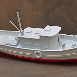 Op de afbeelding: Wit modelbootje met een rode waterlijn. De boot heeft een kajuit, mast en reddingsboei. De boot is een miniatuur replica van een vissersboot.