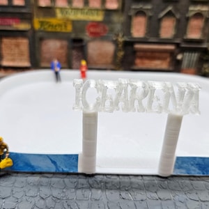 Op de afbeelding: Miniatuur schaatsbaan met een wit oppervlak en blauwe rand. Een bord met de tekst "ICE ARENA" staat op de voorgrond. Kleine beeldjes staan op het ijs en rond de baan, met een achtergrond van een miniatuurstad.