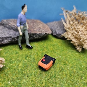 Könnte beinhalten: Miniatur-Mähroboter in Orange und Schwarz mit rotem Knopf, auf einer grünen Oberfläche. Eine kleine Figur in blauer und grauer Kleidung steht in der Nähe von grauen Felsen und getrockneten Pflanzen. Die Szene spielt vor einem blauen Hintergrund.