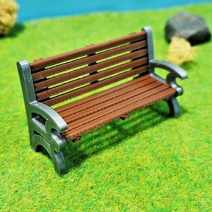 Könnte beinhalten: Eine Miniatur-Parkbank aus Holz mit einer braunen Holzbank und grauen Metallbeinen. Die Bank steht auf einer grünen Grasfläche.