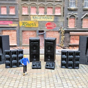 Puede incluir: Un conjunto de sistemas de altavoces negros en miniatura, que incluyen grandes torres, altavoces individuales y matrices más pequeñas. Una figura en miniatura con una chaqueta azul y un casco amarillo está cerca. La escena se desarrolla contra un telón de fondo de un paisaje urbano detallado.