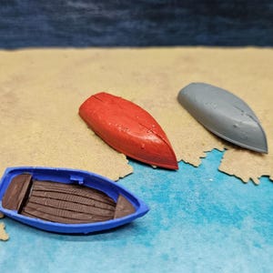 May include: Three miniature toy boats on a sandy surface with a blue background. One boat is blue with a brown interior, and the other two are red and grey. The boats are arranged to suggest a coastal scene.