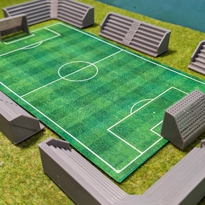 Op de afbeelding: Een miniatuur voetbalveldset met een groen veld, witte markeringen en grijze stadionzitplaatsen. De set bevat doelen en tribunes, ideaal voor tafelspellen of modelpresentaties. Het veld meet ongeveer 30 cm bij 20 cm.