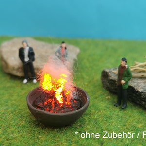 Peut inclure: Un feu de camp miniature avec des flammes orange brûlant dans un foyer en métal noir. Trois figurines miniatures se tiennent autour du foyer sur une surface herbeuse verte. Le texte "ohne Zubehör / Figuren" est visible en bas de l'image.