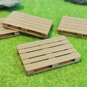 Könnte beinhalten: Vier kleine, hellbraune Holzpaletten liegen auf einer grünen Oberfläche. Die Paletten sind rechteckig mit einer Lattenoberseite und einem Zwei-Wege-Eingang. Der Hintergrund besteht aus grünem Gras und kleinen Steinen.