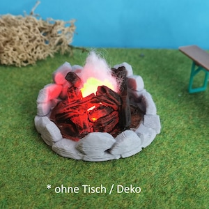 May include: A miniature campfire with glowing red and orange embers, surrounded by grey rocks, on a green grassy surface. A brown wooden picnic table with green metal legs is in the background.