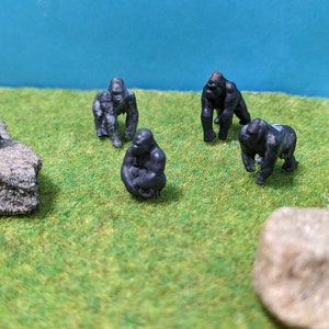 Op de afbeelding: Een set van zes zwarte gorilla-figuurtjes, sommige zittend en sommige staand, op een groene grasmat met twee grijze stenen.