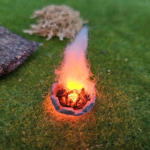 May include: A miniature campfire with bright orange and yellow flames, set in a small, grey, stone-like bowl. The scene is on a green, grassy surface, with two dark grey rocks and a clump of tan material nearby. A fantasy or tabletop gaming item.