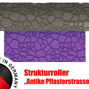 Puede incluir: Un rodillo texturizado morado con un patrón de adoquines. El rodillo está etiquetado como "Strukturroller "Antik Pflastersteinstrasse" y "Made in Germany".