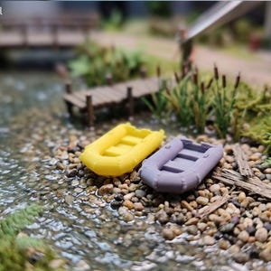 Könnte beinhalten: Miniatur-Szene mit einem gelben und einem lila Schlauchboot, die an einem kiesigen Flussufer liegen. Die Boote befinden sich neben einem kleinen Holzsteg und einem fließenden Bach. Die Szene ist von Grün umgeben.