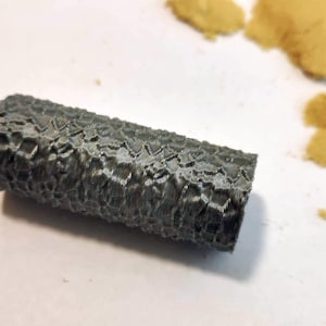 May include: A grey, textured, cylindrical object with a rough surface. The object is on a white surface with a pile of yellow sand in the background.