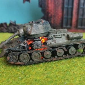May include: A toy tank model painted in green and grey. The tank is on fire with orange flames coming from the inside. The tank is sitting on a green grassy surface.